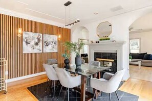 Un espace repas comprenant une table en verre, des chaises blanches, un mur avec des lambris en bois et une cheminée.
