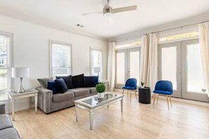 Un salon moderne avec un canapé, une table basse et des fauteuils, de grandes fenêtres avec des rideaux et un ventilateur de plafond.