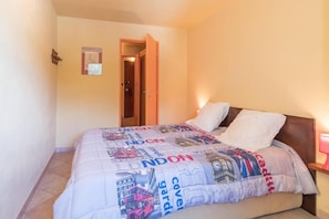 Cama com colcha decorada com tema urbano, cabeceira de madeira, abajur e porta que dá para outro quarto.