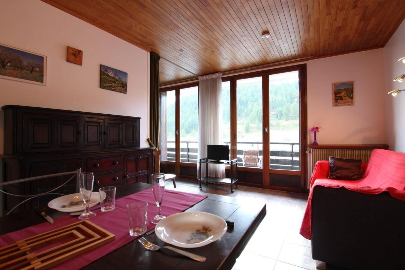A dining area with a wooden table set for two, a red sofa, and large windows offering a view of the outdoors.