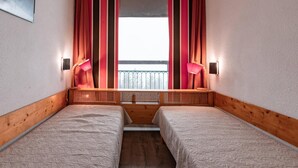 A small, cozy room with two beds, a window with striped curtains, and two wall-mounted lamps.