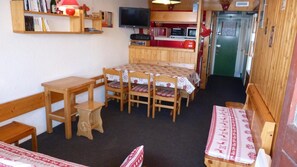 A compact room with a dining table, chairs, a small kitchen area with a microwave and toaster, and a television on the wall.