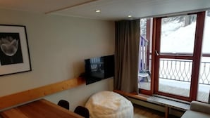 Une chambre moderne avec un banc en bois, un fauteuil en forme de poire blanc, un téléviseur à écran plat et une grande fenêtre donnant sur un paysage enneigé.
