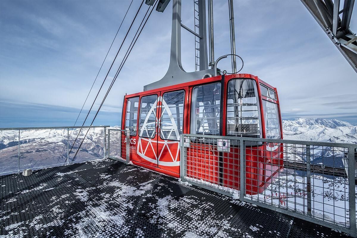 Une télécabine rouge, dotée d’une structure métallique et de fenêtres en verre, suspendue par des câbles devant un paysage montagneux enneigé.