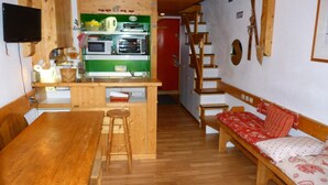 Een compacte keuken met een houten tafel en krukken, een klein barretje en een rode deur die leidt naar een trap.