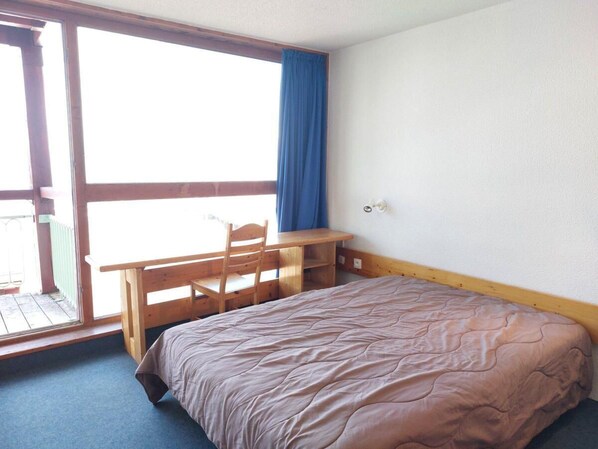 Une chambre à coucher avec un lit, un bureau en bois et une chaise. Il y a une fenêtre donnant sur un balcon.
