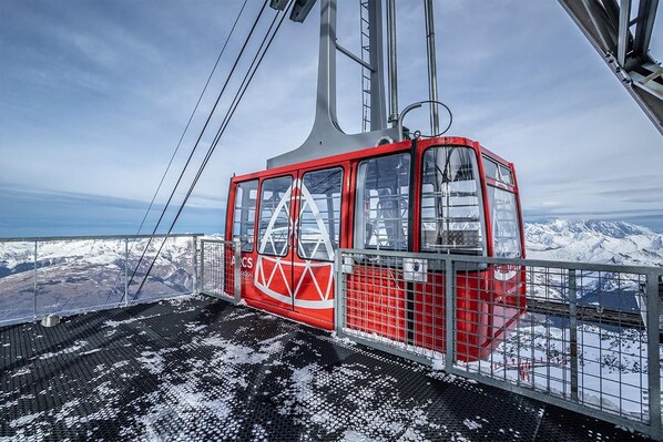 A red cable car with a metal frame and glass windows, suspended by cables against a snowy mountain backdrop.