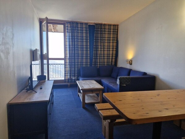 A small, sparsely furnished room with a sofa, a wooden table, and a bench. There is a window with curtains, a radiator, and a wall-mounted light fixture.