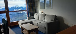 A living room with a sofa, a small wooden table, and a view of snow-capped mountains through a balcony with a metal railing.