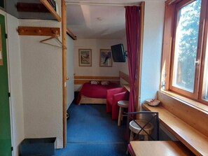 Ein kleines Zimmer mit Bett, Tisch, Stuhl und einem Fenster mit Vorhängen.