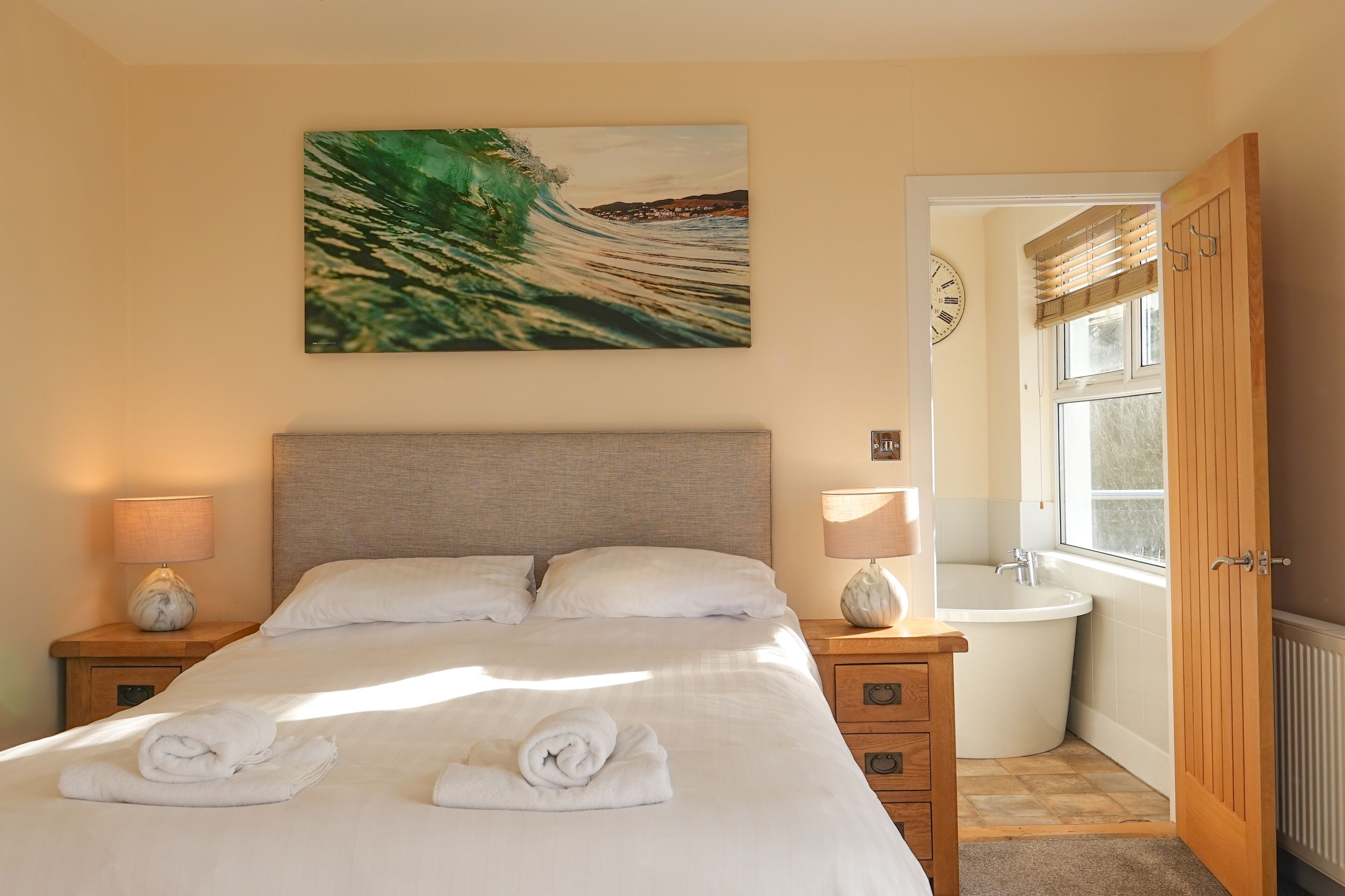 A neatly made bed with towels, a painting of a wave, a wooden nightstand, a lamp, a bathroom with a bathtub, and a wooden door.