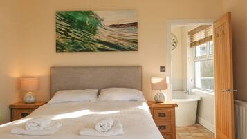A neatly made bed with towels, a painting of a wave, a wooden nightstand, a lamp, a bathroom with a bathtub, and a wooden door.