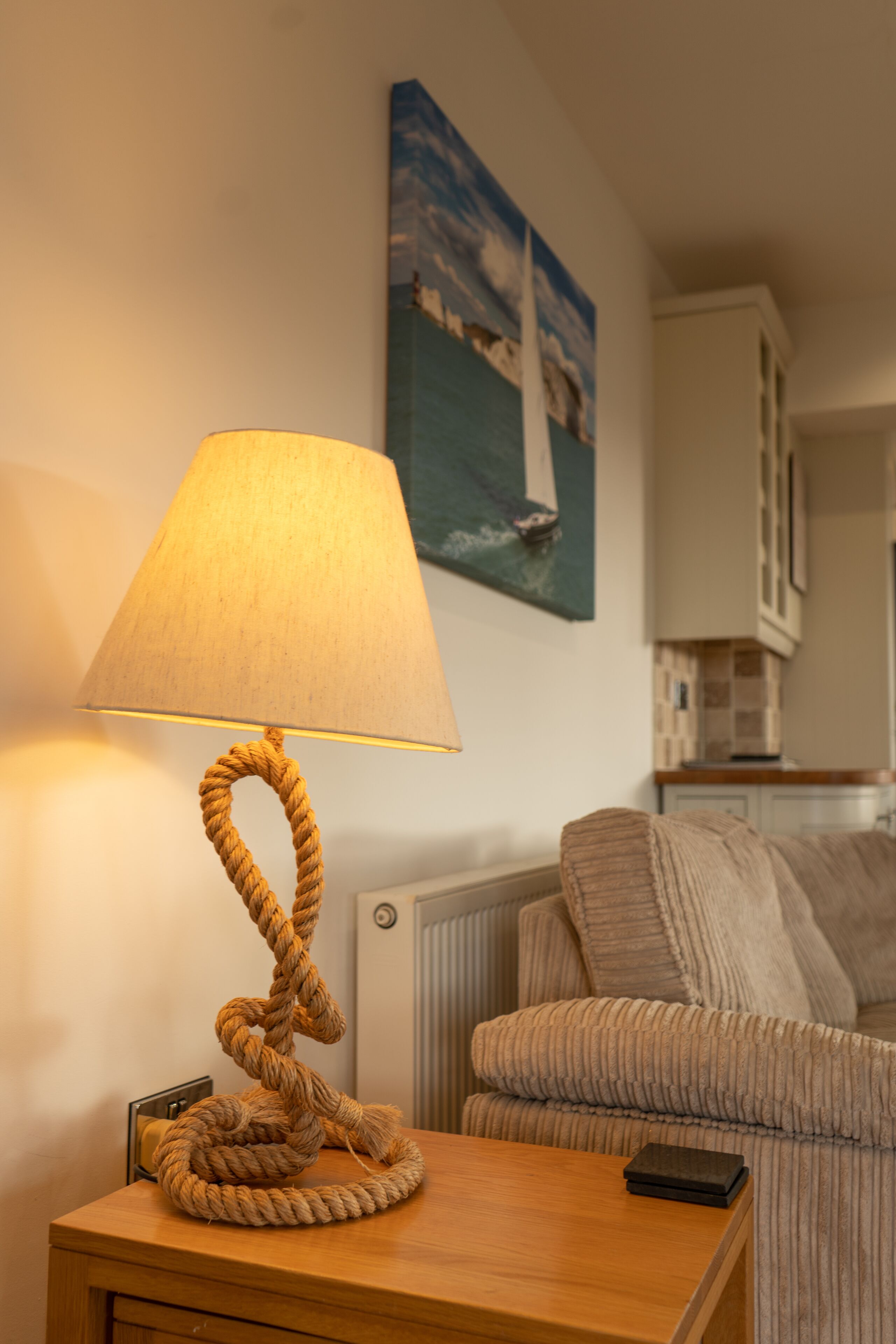 Una mesita de noche de madera con una lámpara de cuerda, un sofá a rayas y un cuadro de un velero en la pared.