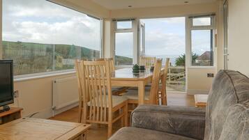 A room with a dining table and chairs, a sofa, a TV, and large windows offering a view of the outdoors.