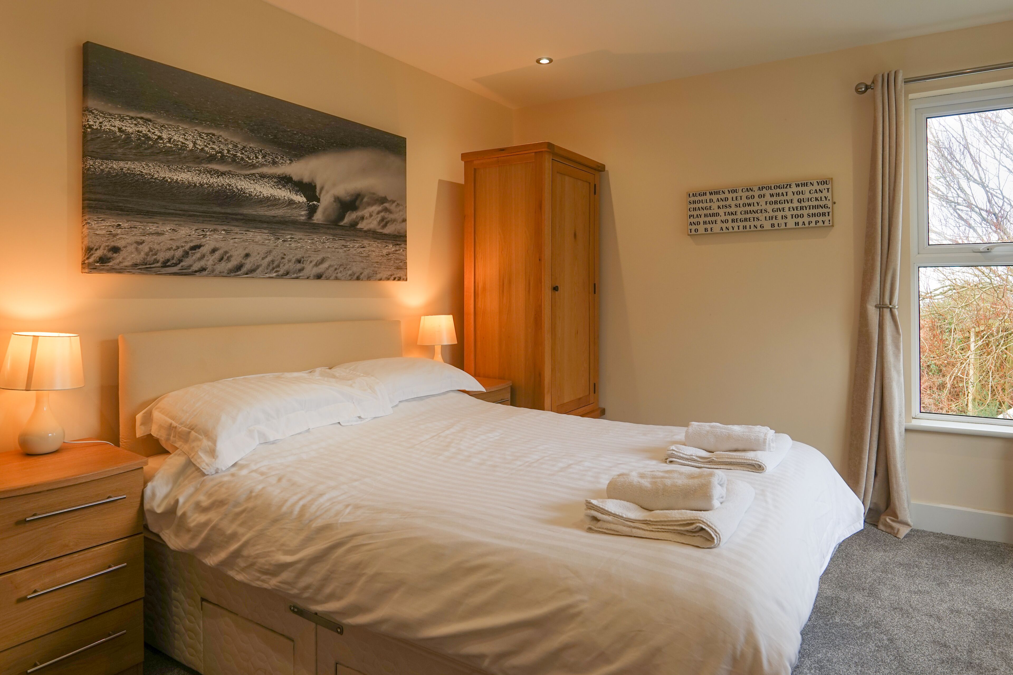 A neatly made bed with white linens and towels, a wooden wardrobe, a wall-mounted painting of a wave, and a motivational quote on the wall.