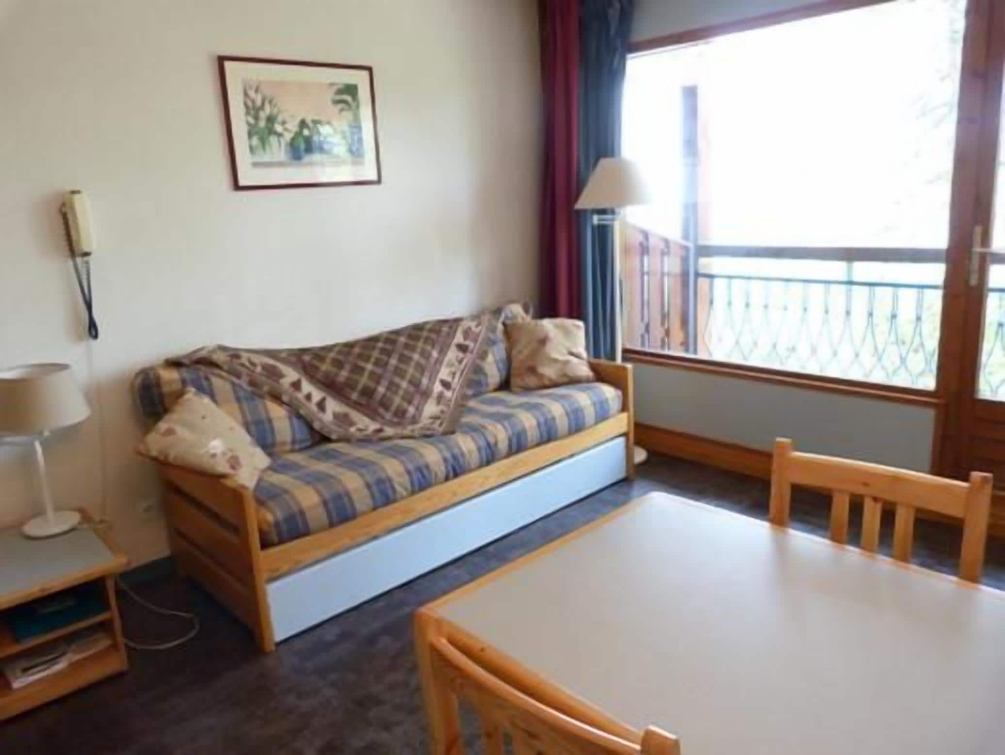 A small, cozy room with a sofa, a table, and a chair. There is a window with a view of a balcony.