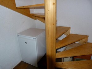 A compact white refrigerator in a corner, wooden stairs leading to a lower level, and a white wall.