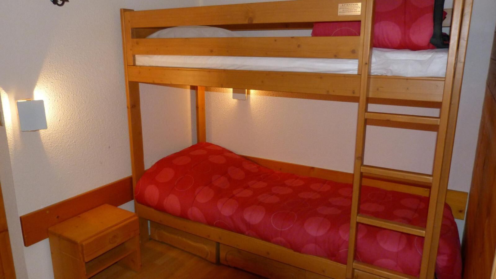 A bunk bed with a red patterned mattress, a wooden bench, and a wall-mounted light fixture.