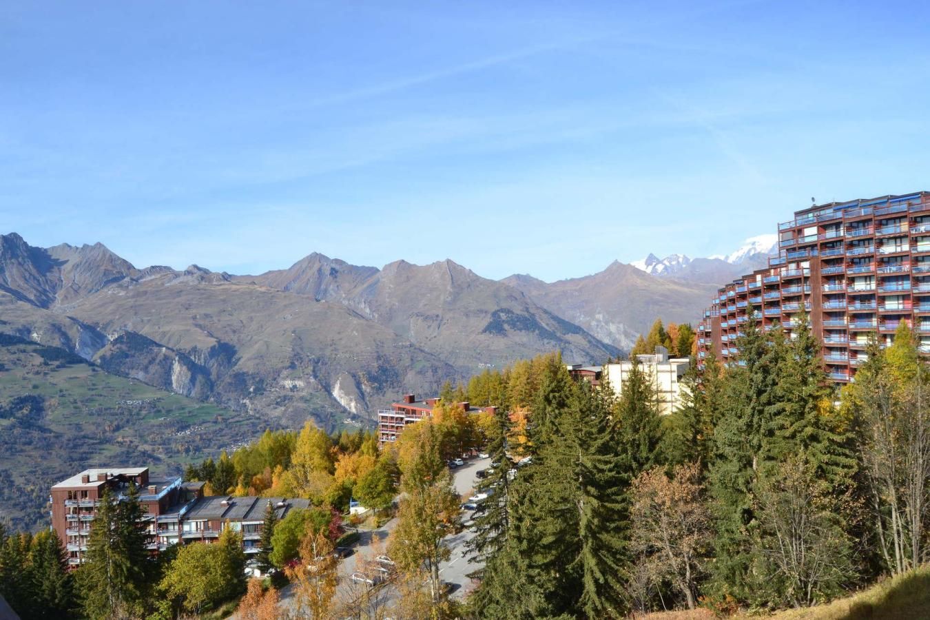 A mountainous landscape with a hotel featuring multiple floors and balconies, surrounded by trees and a clear sky.