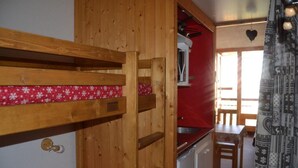 Een houten stapelbed met een matras met een rood-wit patroon, een kitchenette met een spoelbak en een koelkast, en een gordijn met een dessin.