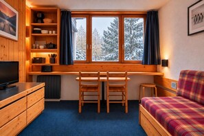 A cozy cabin room with a wooden desk, two chairs, a television, a sofa, and a window with a snowy view.