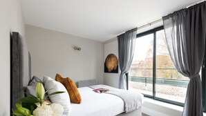 A modern bedroom with a large window, a bed with white bedding, a gray headboard, and a decorative wall piece. - Fitzrovia apartments by DMONTRIO (London)