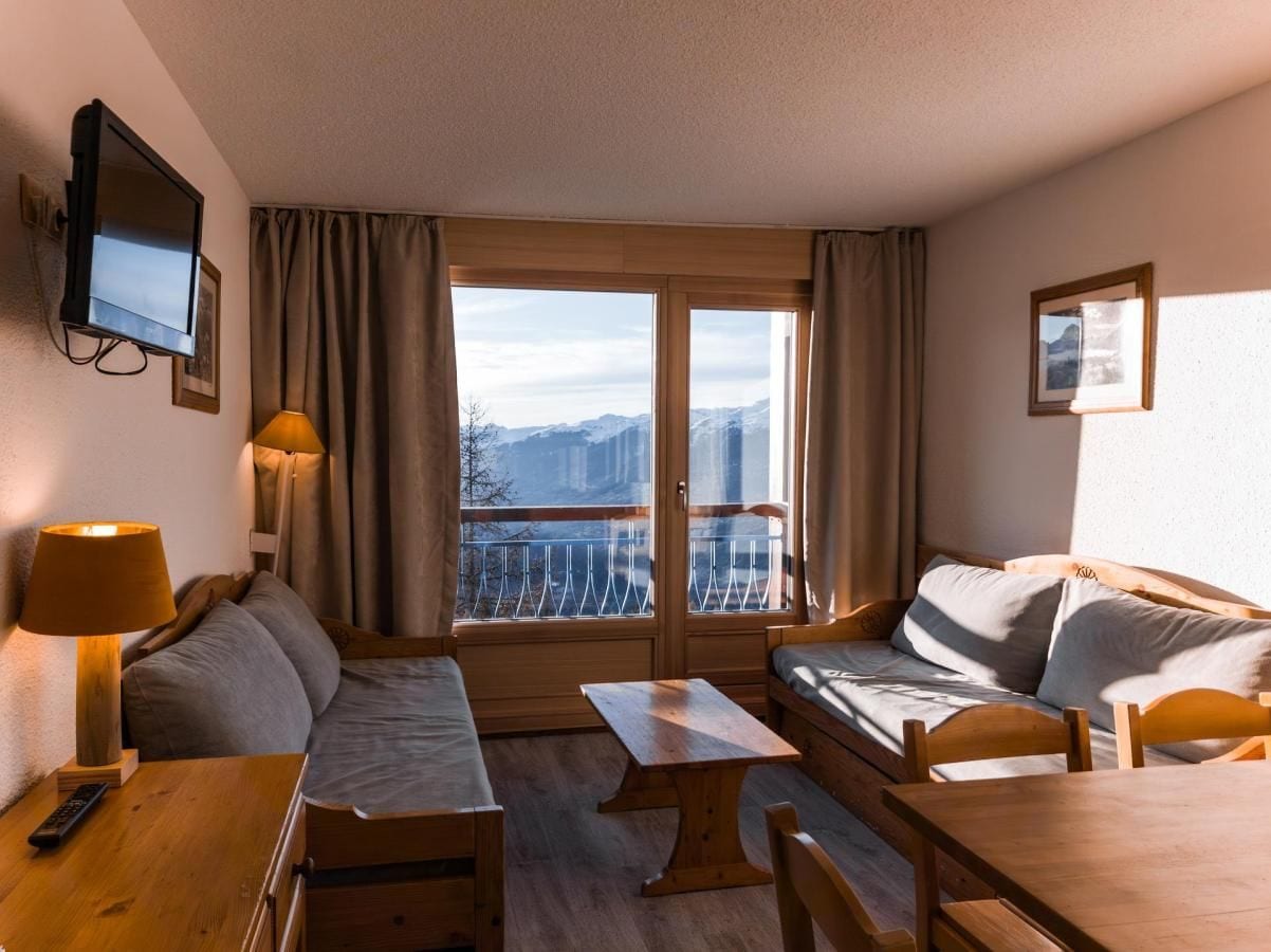 A cozy hotel room with a sofa, a small table, a TV mounted on the wall, and a window with a mountain view.