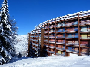 A multi-story hotel with balconies, surrounded by snow-covered trees and a clear blue sky.