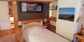 Quarto com cama, televisão na parede, prateleira com objetos decorativos e quadro pendurado na parede.