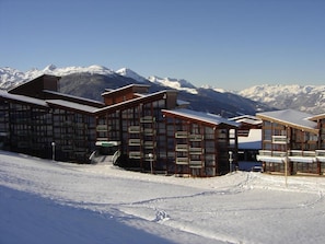 Ein schneebedecktes Skigebiet mit Holzhäusern, blauem Himmel und verschneiten Bergen im Hintergrund.