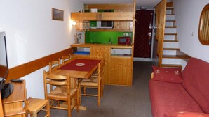 Un espace de vie compact comprenant une table à manger en bois, des chaises, un canapé rouge, un coin cuisine avec un micro-ondes et une étagère verte, ainsi qu’un escalier en bois.