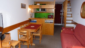 A compact living space with a wooden dining table, chairs, a red sofa, a kitchen area with a microwave and a green shelf, and a wooden staircase.