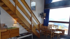 Una habitación con una escalera de madera, una zona de comedor con sillas y una mesa de madera, un pequeño banco blanco y vistas de montañas a través de una ventana.