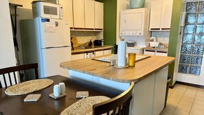 Une cuisine moderne avec des armoires blanches, un four à micro-ondes, une cuisinière et un évier. On y trouve un îlot central en bois, doté d’un tabouret de bar et d’un set de table en fibres tressées.