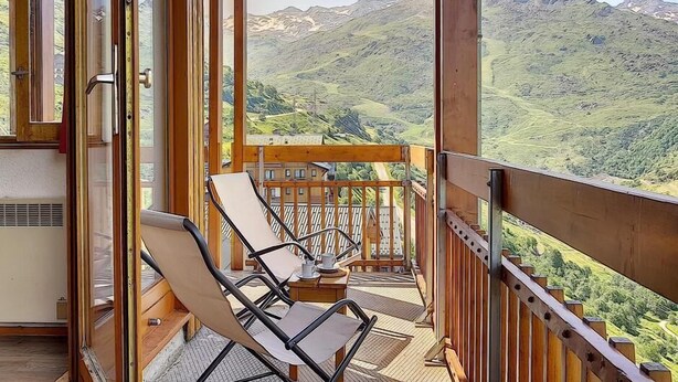 Un balcón con vista a un paisaje montañoso, baranda de madera y dos sillones de relax.