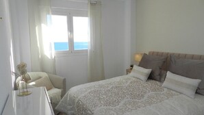 Ein Schlafzimmer mit Bett, Nachttisch, Stuhl, einem Fenster mit Vorhängen und Meerblick.