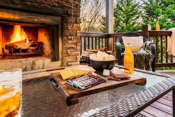 A cozy outdoor seating area with a stone fireplace, a glass table, snacks, and a bottle of Crush orange.