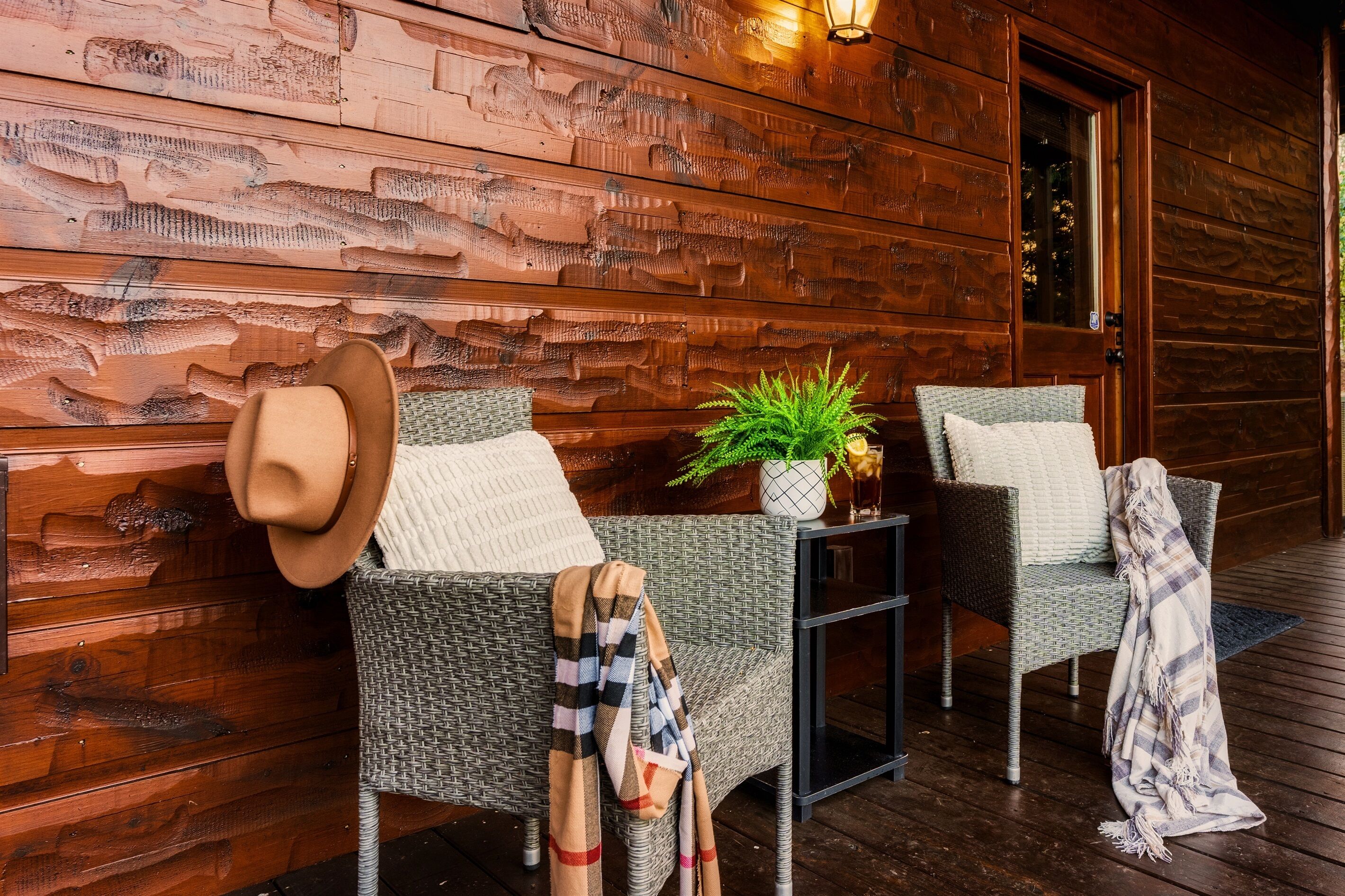 A wooden exterior wall with a textured pattern, two wicker chairs with cushions, a small table with a plant, and a door.