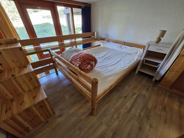 A wooden bunk bed with a mattress and pillows, a bedside table with a lamp, and a window with curtains.