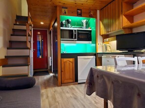A compact kitchen with a microwave, a small refrigerator, and wooden cabinets. A dining table with a tablecloth is visible in the foreground.
