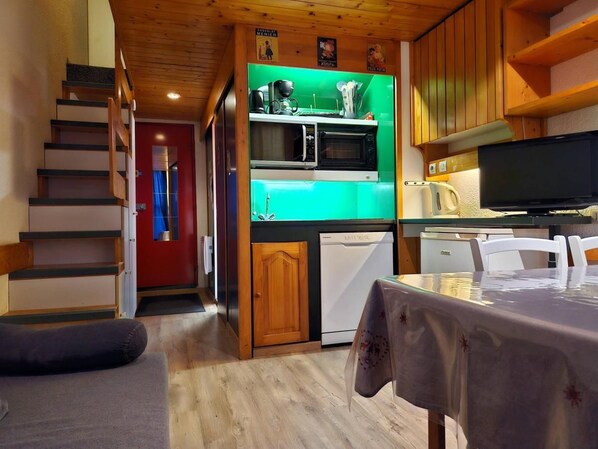 Une cuisine compacte équipée d’un four à micro-ondes, d’un petit réfrigérateur et de meubles de rangement en bois. Une table à manger recouverte d’une nappe est visible au premier plan.