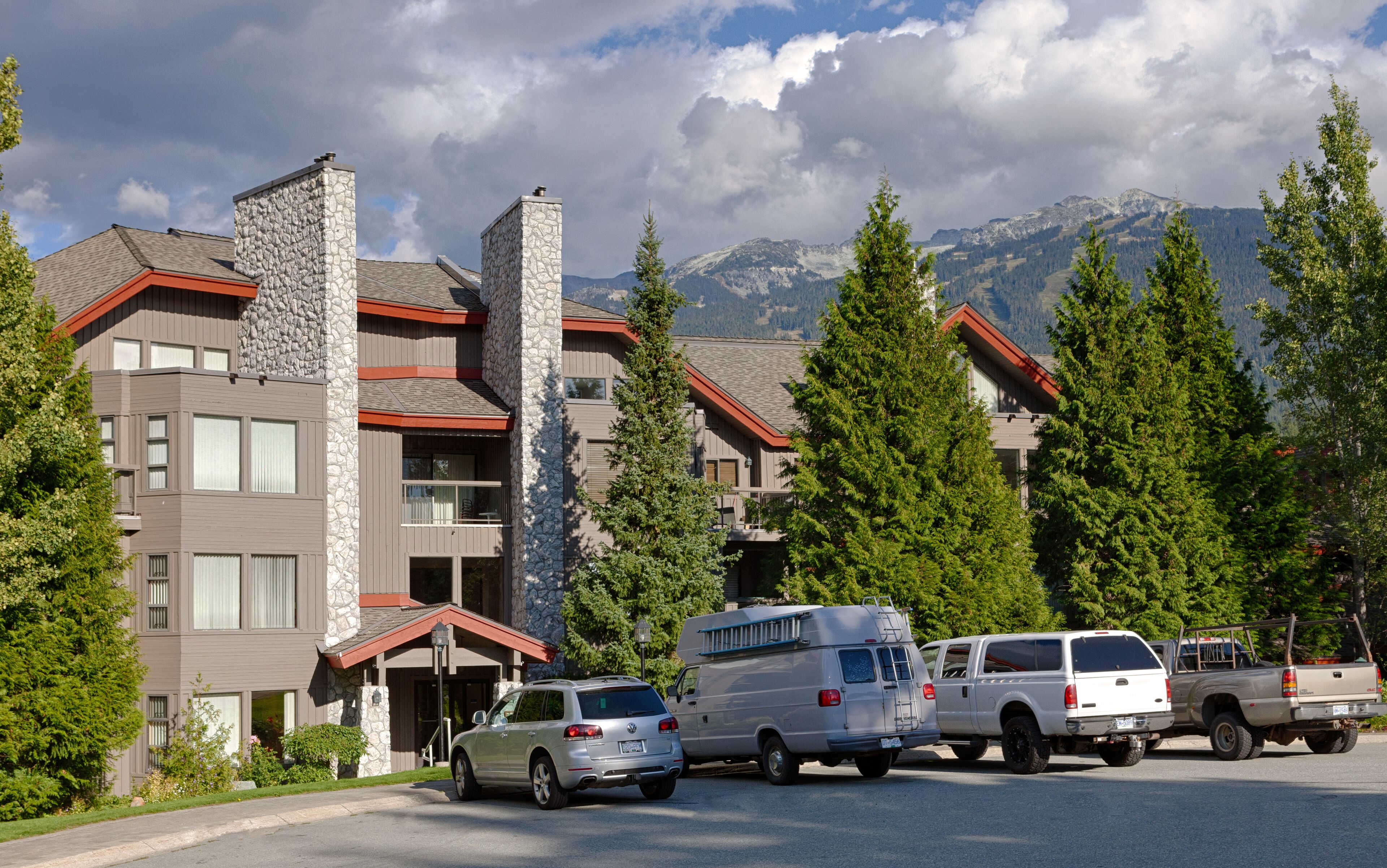 A multi-story building with stone facades and red roofs, surrounded by tall trees and mountains in the background.
