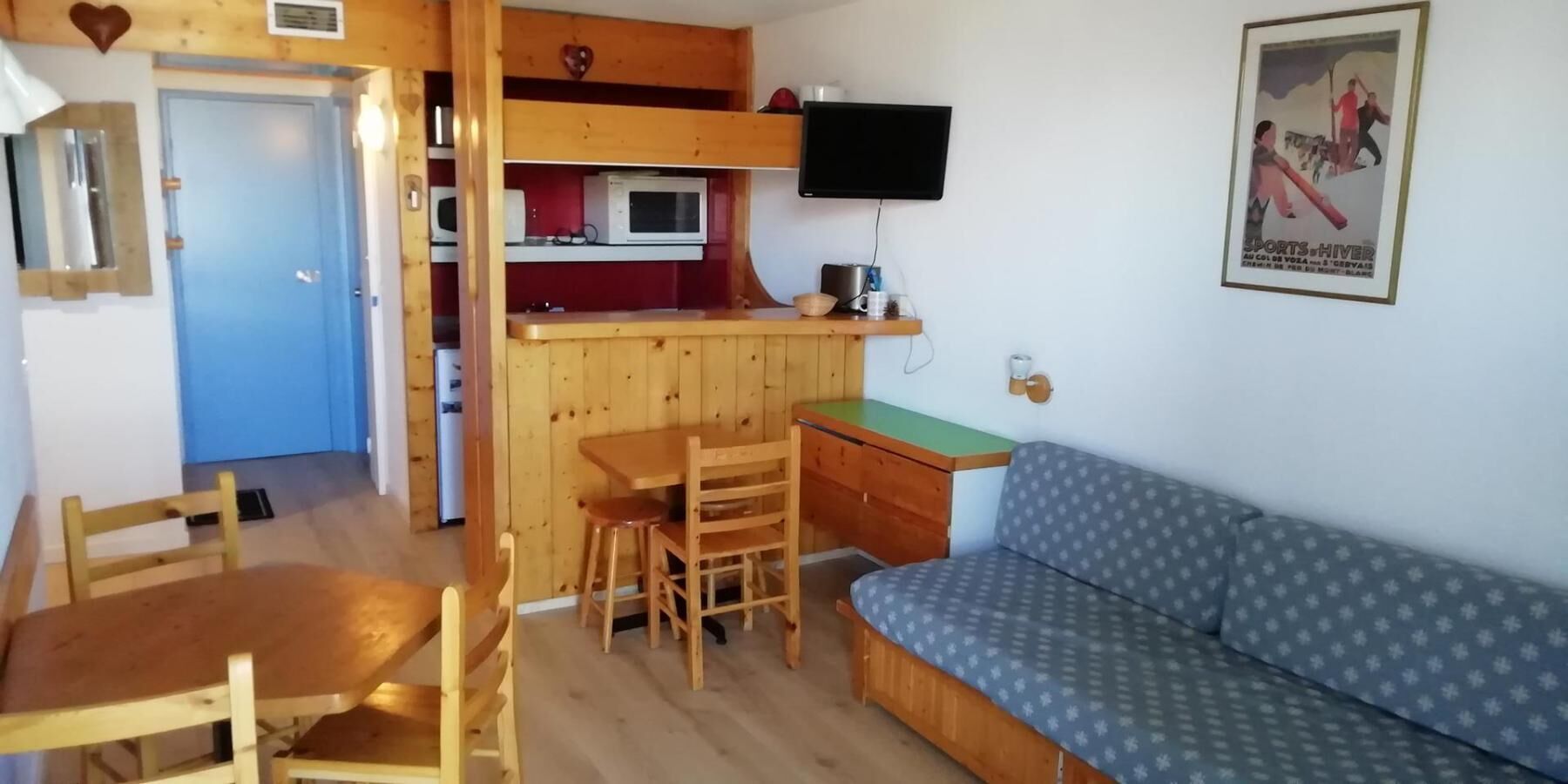 Une pièce compacte comprenant un coin cuisine, une table à manger en bois avec des chaises, un canapé et un téléviseur fixé au mur.