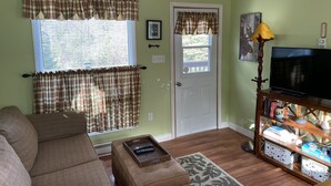A living room with a couch, a television on a wooden stand, a door with a window, and a wall with a framed picture. - Hilltop Cottage steps from East Coast Trail, fishing & whale watching (Cape Broyle)