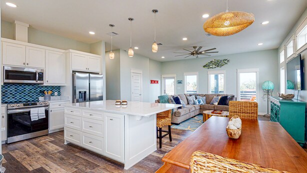 A modern kitchen with white cabinetry, a central island, and a dining area with a wooden table. - Heated Private Pool, Yard Games, Golf Cart Included (Port Aransas)