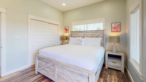 A bedroom with a wooden bed frame, white bedding, a nightstand with a lamp, and a window with blinds. - Heated Private Pool, Yard Games, Golf Cart Included (Port Aransas)