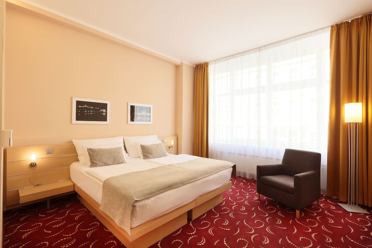 Habitación de hotel con una cama grande, una silla, una mesita de noche y un ventanal con cortinas.