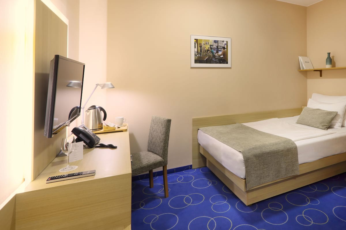Habitación de hotel con cama, escritorio con teléfono, silla, cuadro en la pared y ventana con cortina ligera.