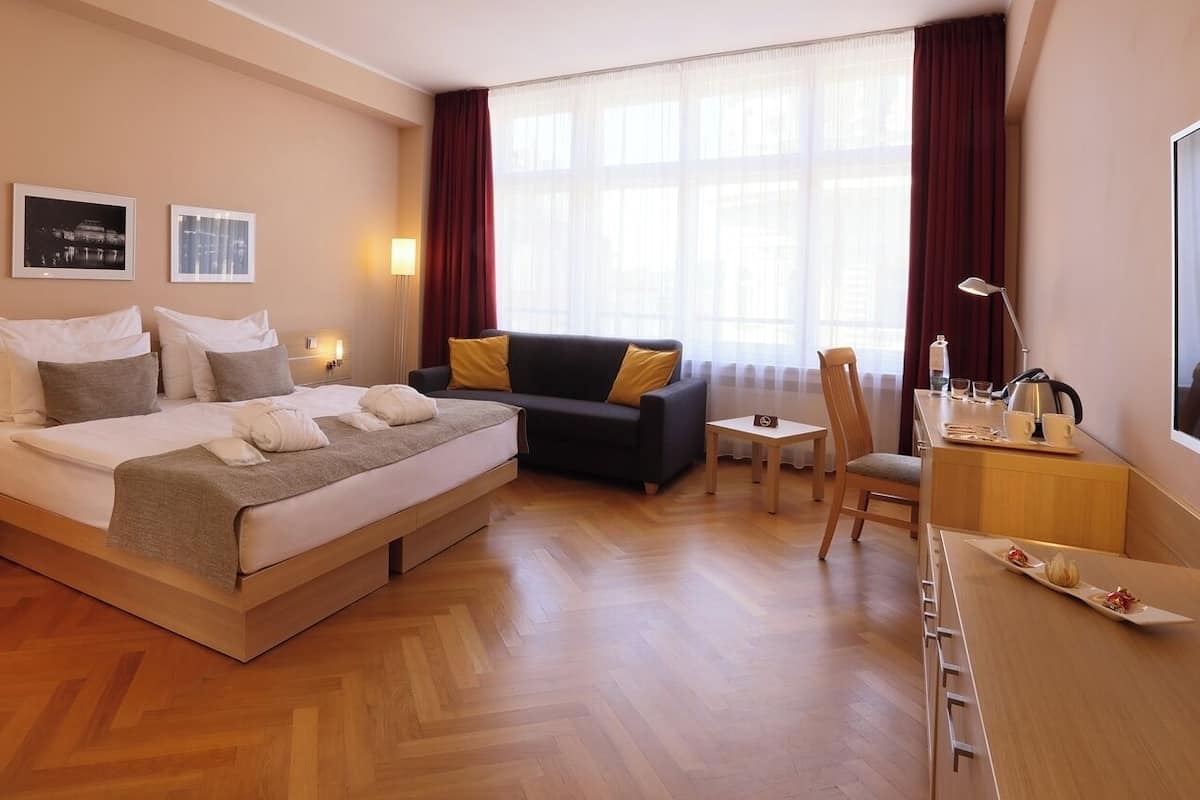 Habitación de hotel con cama, sofá, escritorio y silla. Se observan dos cuadros en la pared, una lámpara y una ventana con cortinas.
