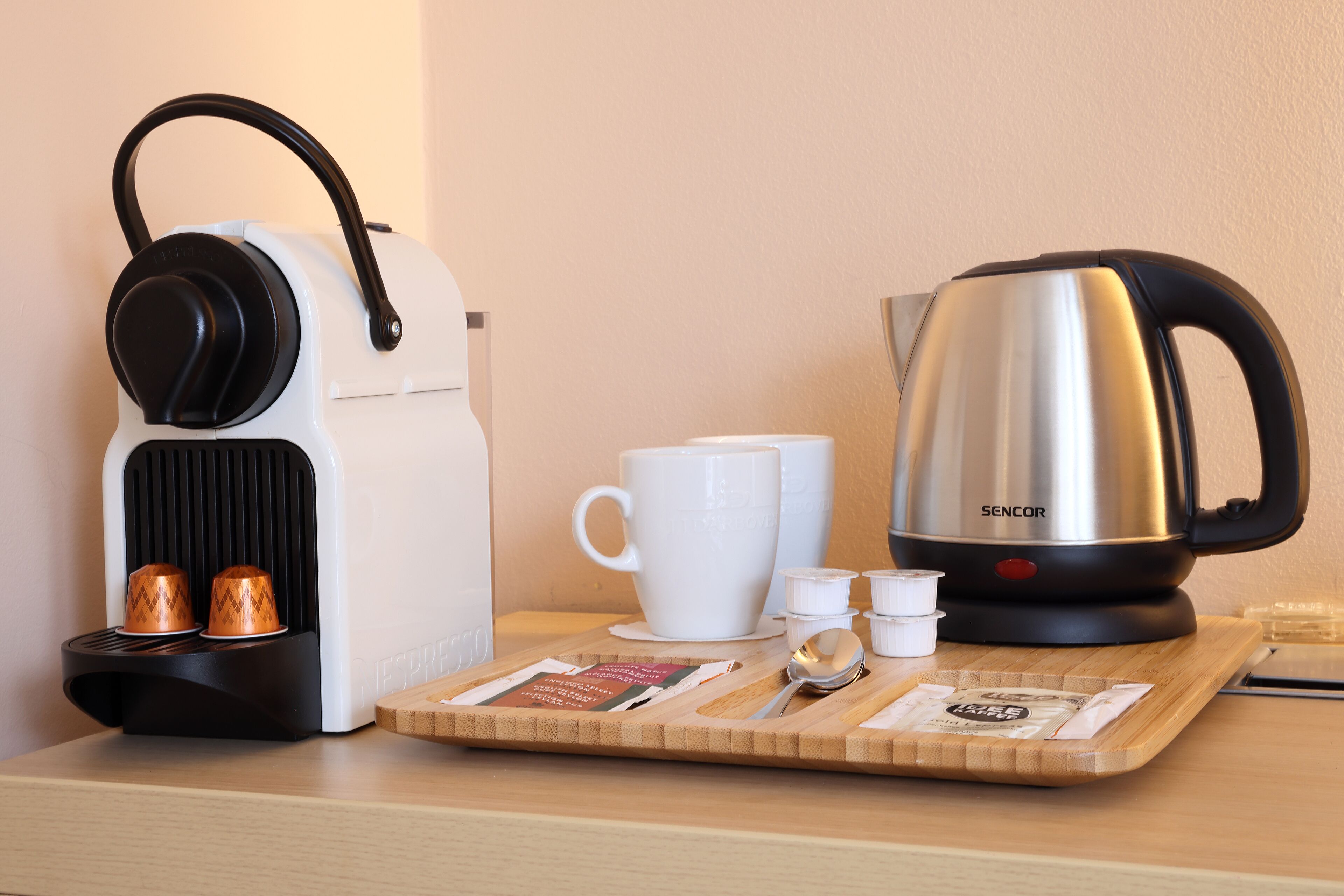 A coffee maker, a cup, a kettle, and a tray with coffee pods and a spoon on a wooden table.
